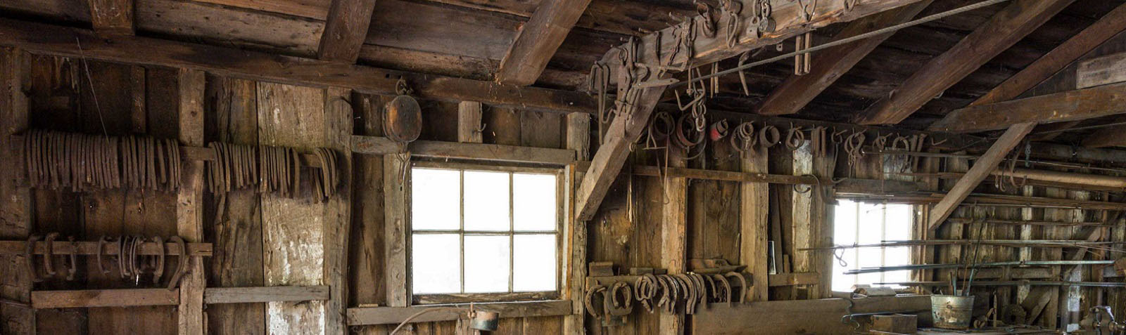Photo couleur de l'intérieur d'une forge. Des outils de forge sont accrochés le long du mur et sur les poutres du plafond.